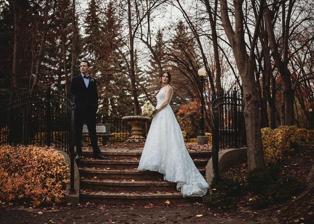 Bride in a flowing white gown and groom in a black suit pose on stone steps in an autumn garden with bare trees and golden foliage.