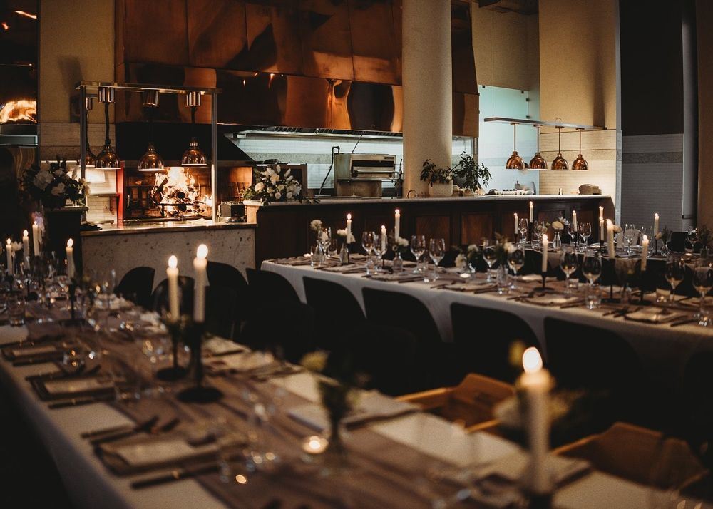 Candlelit restaurant dining room with long set tables, elegant place settings, and a warm open kitchen glowing in the background.