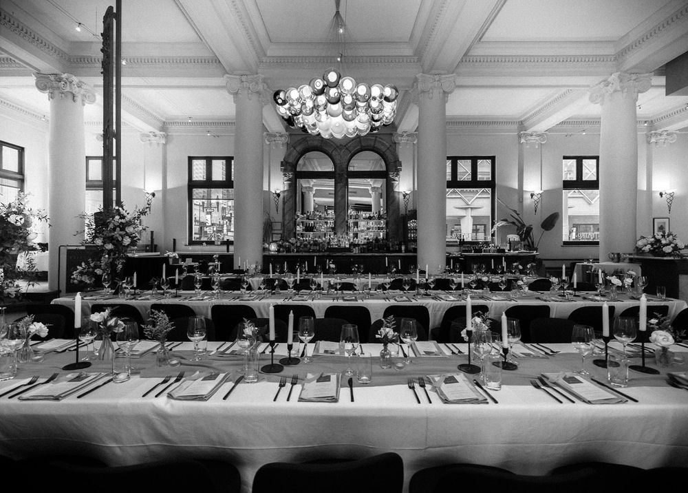 Elegant black and white view of a grand banquet hall with long set tables, floral centerpieces, ornate columns and a chandelier.