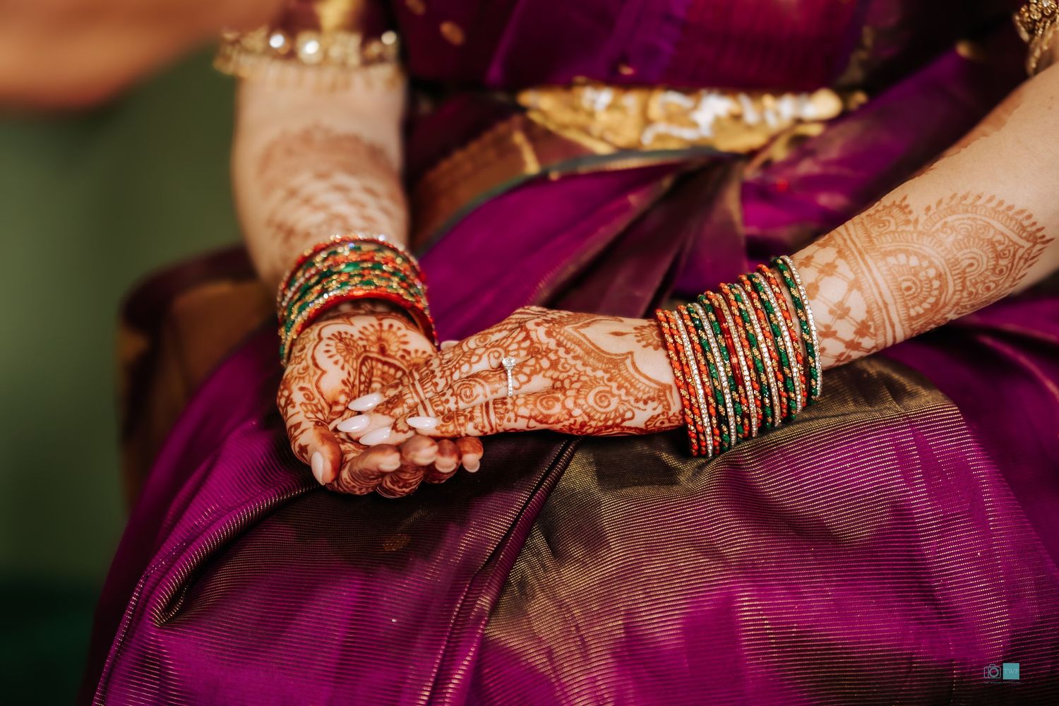 Bride's hands with intricate henna mehndi designs and green and red bangles rest on a rich purple silk saree.