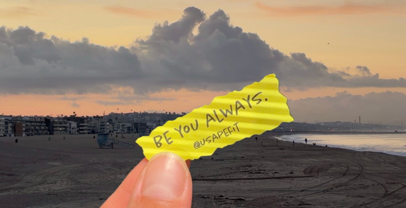 Hand holding a yellow wrapper reading "Be you always" in front of a beach at sunset with dramatic clouds over the shoreline.