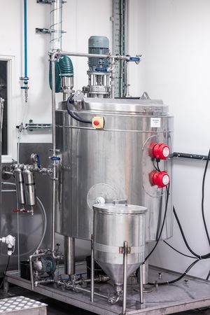 Stainless steel industrial mixing tank with motor, red controls, and piping in a clean facility.
