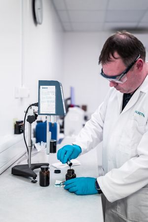 Lab technician in white coat and blue gloves examining chemical samples in a professional laboratory setting.
