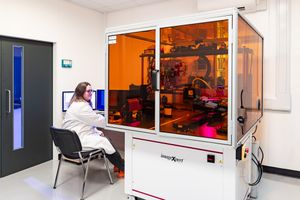 Female scientist in white lab coat working at computer beside large orange-windowed laboratory analysis machine.
