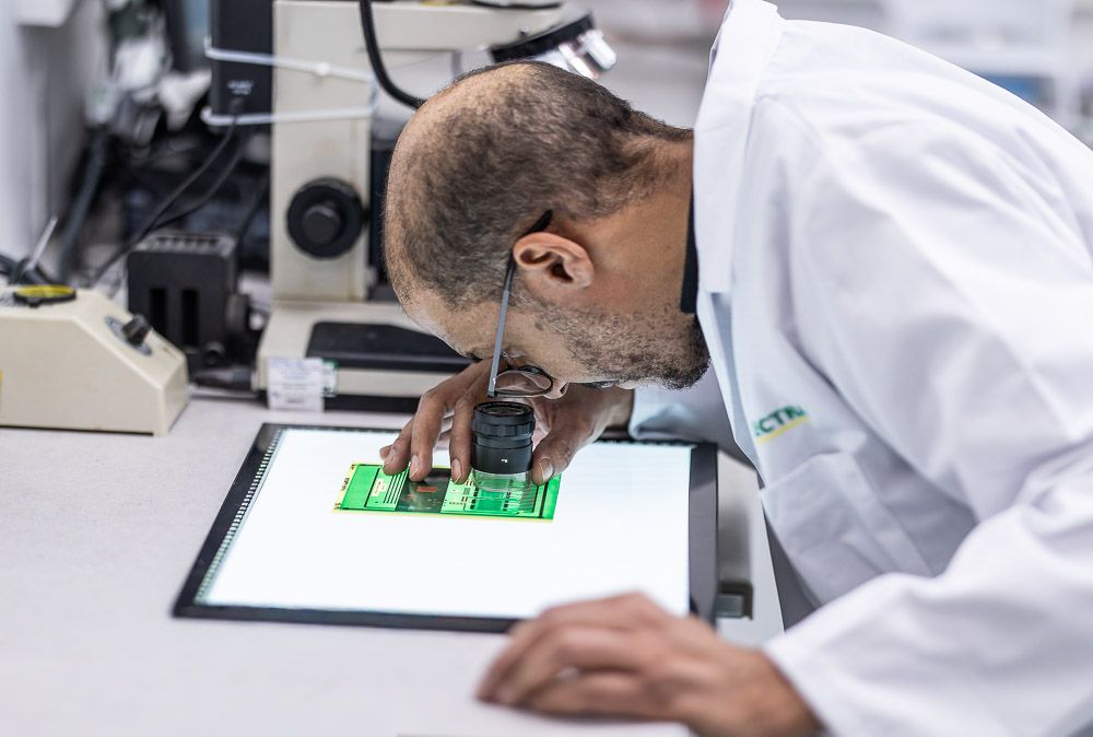 Lab technician in white coat inspecting green circuit board under microscope in professional laboratory setting.