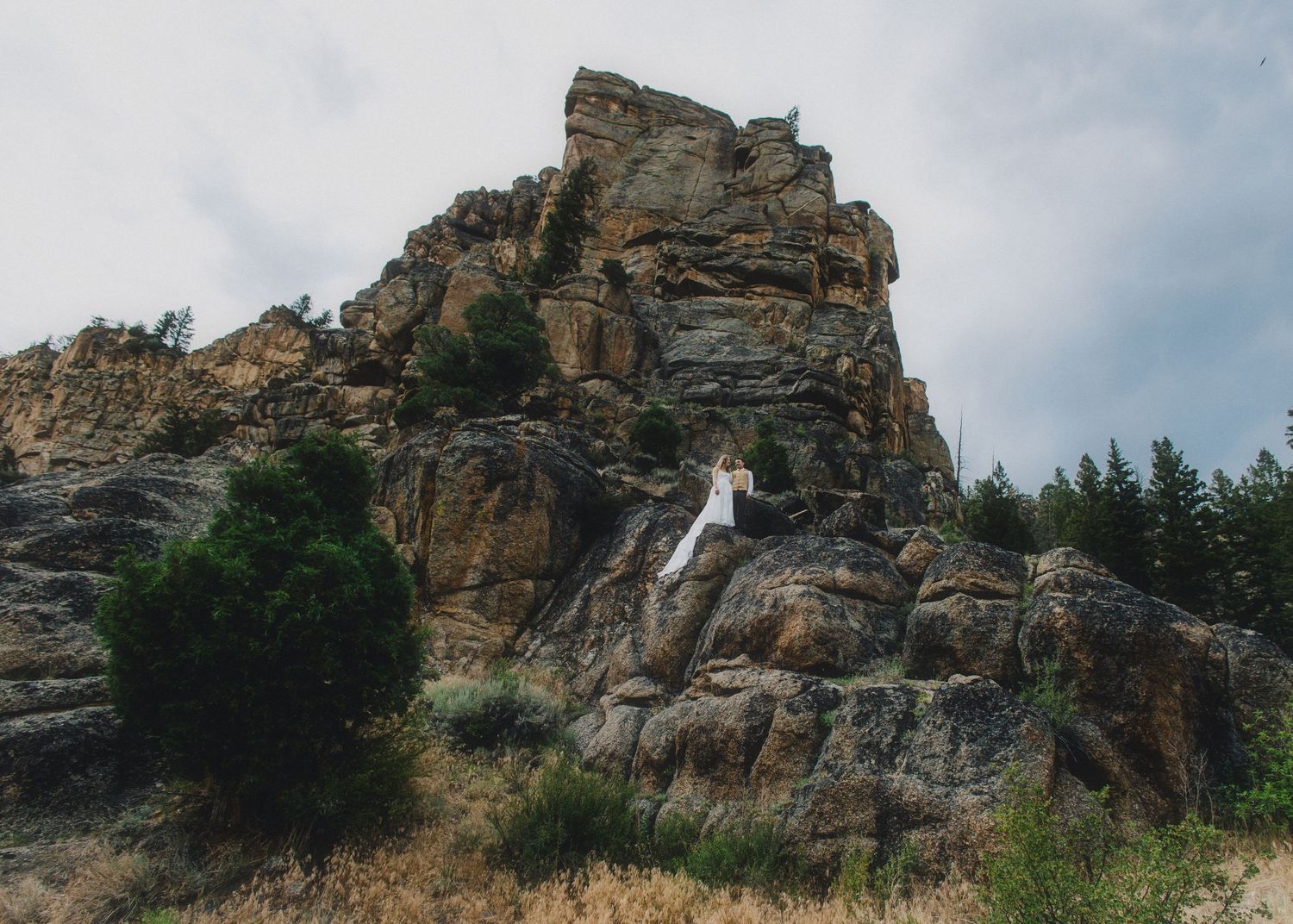 A person in white stands on rugged rocky cliffs surrounded by trees under a cloudy sky.