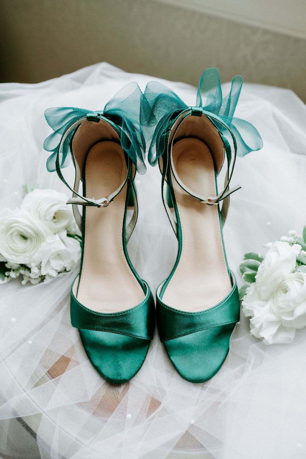 Elegant emerald green satin wedding heels with organza ribbon ankle ties displayed on white bridal veil with white floral bouquets.