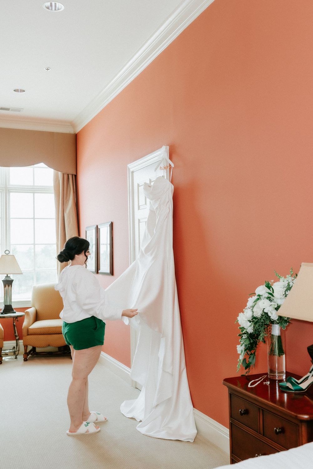 Bride in green shorts holds white wedding gown against coral wall in elegant hotel room before ceremony.