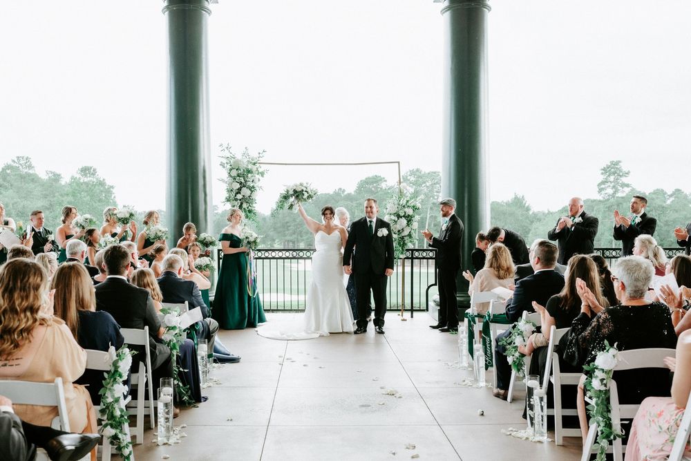 Elegant outdoor wedding ceremony on a columned pavilion with guests seated in white chairs and floral arch backdrop.