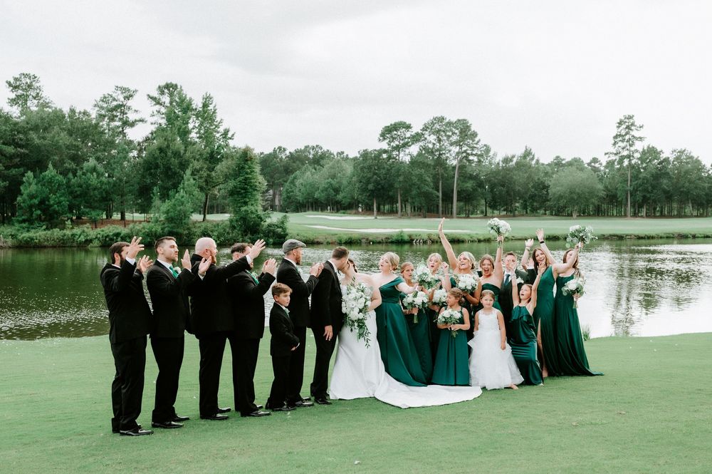 Wedding party in emerald green dresses and black suits celebrating outdoors on a golf course with a lake backdrop.