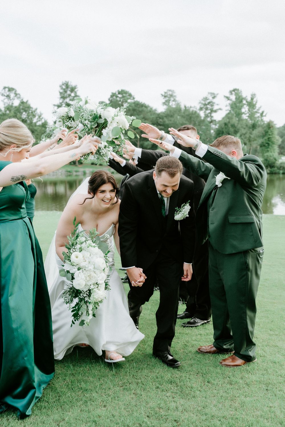 Bride and groom walk through arch of raised hands formed by wedding party in emerald green and black attire outdoors.