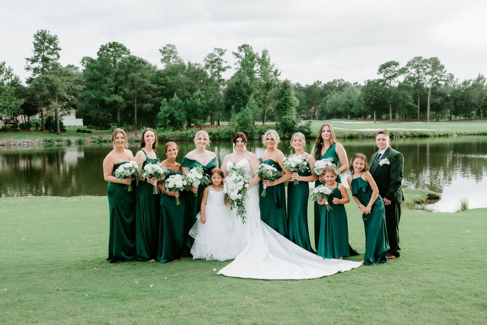 Bride in white gown with bridesmaids in emerald green dresses holding white bouquets outdoors near a pond.