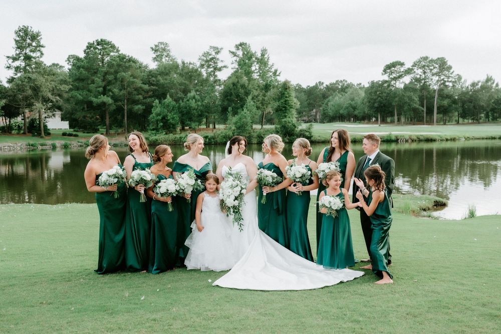 Bride in white gown with bridesmaids in emerald green dresses holding white bouquets on golf course lawn near pond.