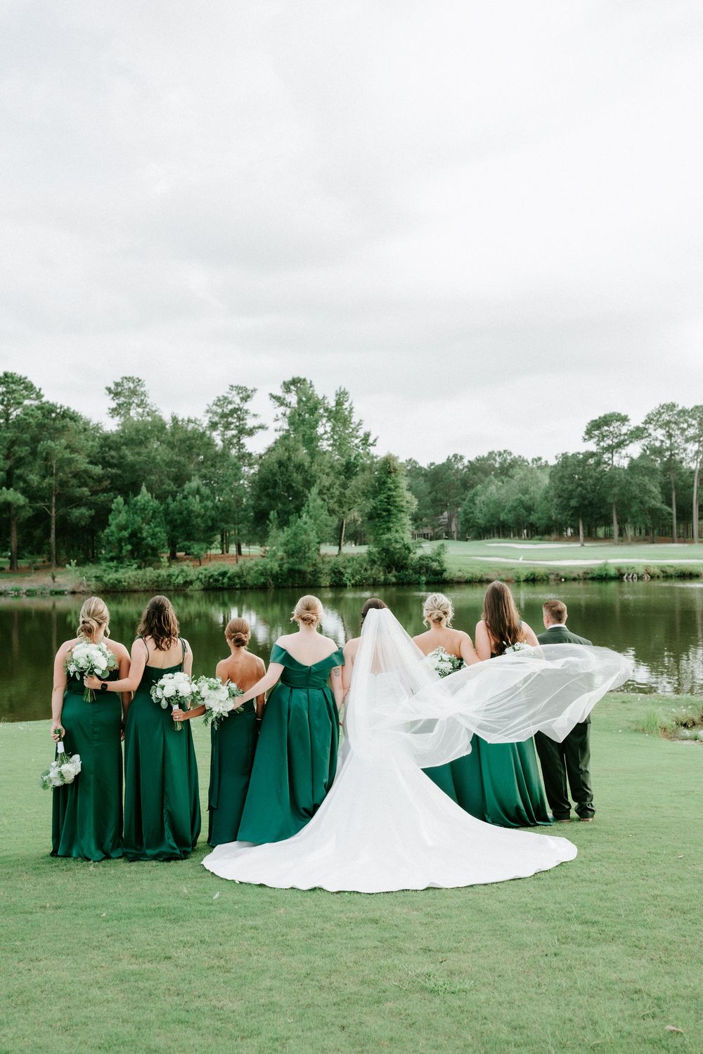 Bridesmaids in emerald green gowns stand with bride in white wedding dress and flowing veil by a serene pond.