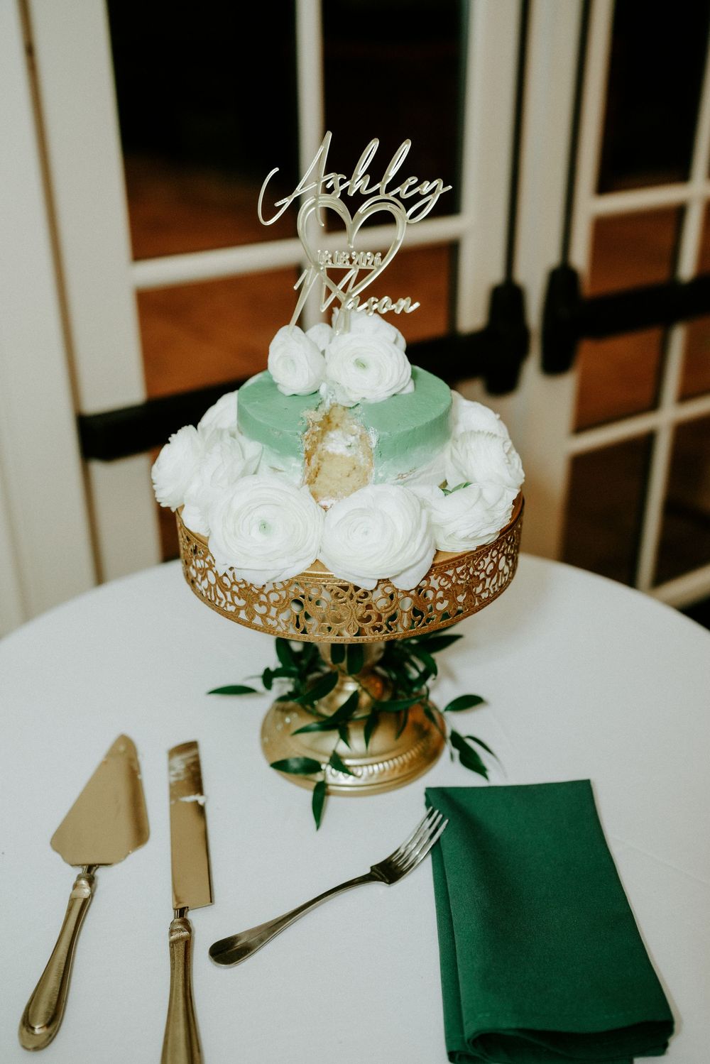 Elegant two-tier mint green wedding cake with white roses on gold stand, personalized topper reading Ashley and Mason.