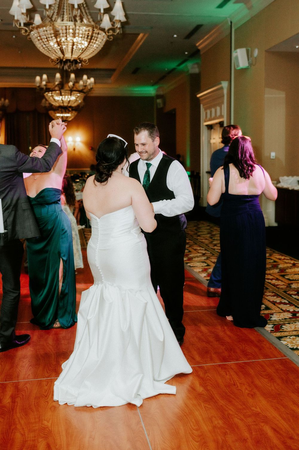 Bride and groom share a first dance in elegant ballroom with crystal chandeliers and green ambient lighting.