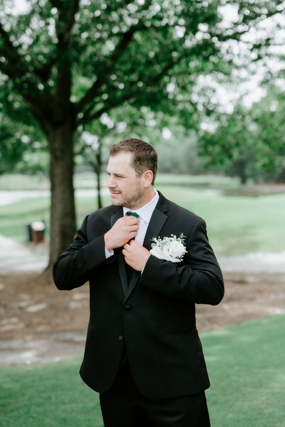 Handsome groom in black tuxedo adjusting bow tie with white floral boutonniere in lush green outdoor setting.