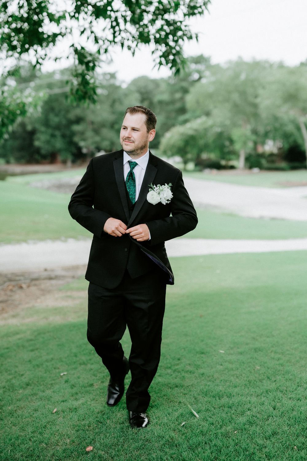 Handsome groom in black tuxedo with green tie and white boutonniere poses outdoors on lush golf course.