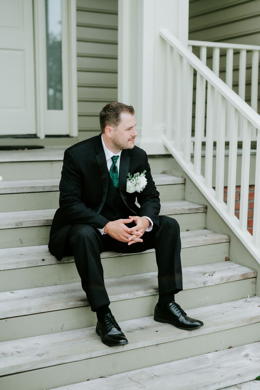 Dapper groom in black suit with emerald green tie and white boutonniere sitting on wooden porch steps.