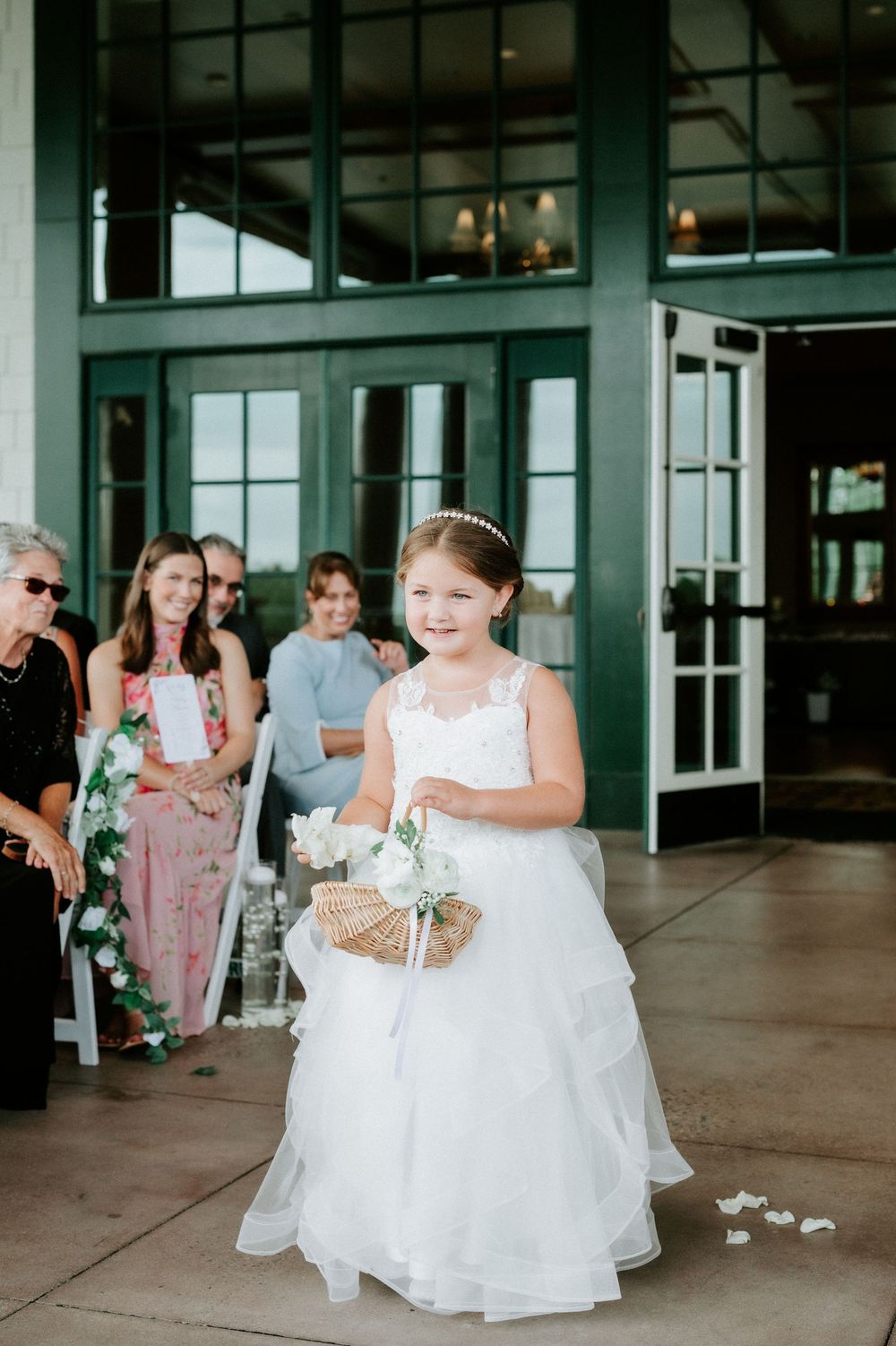 Adorable flower girl in white lace dress carries wicker basket while walking down outdoor wedding aisle.