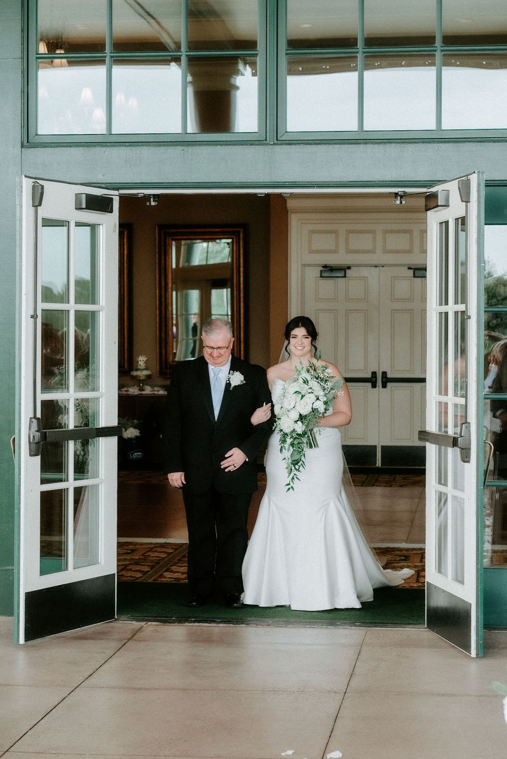Bride in white mermaid gown holding bouquet walks through elegant venue doors escorted by father in tuxedo.