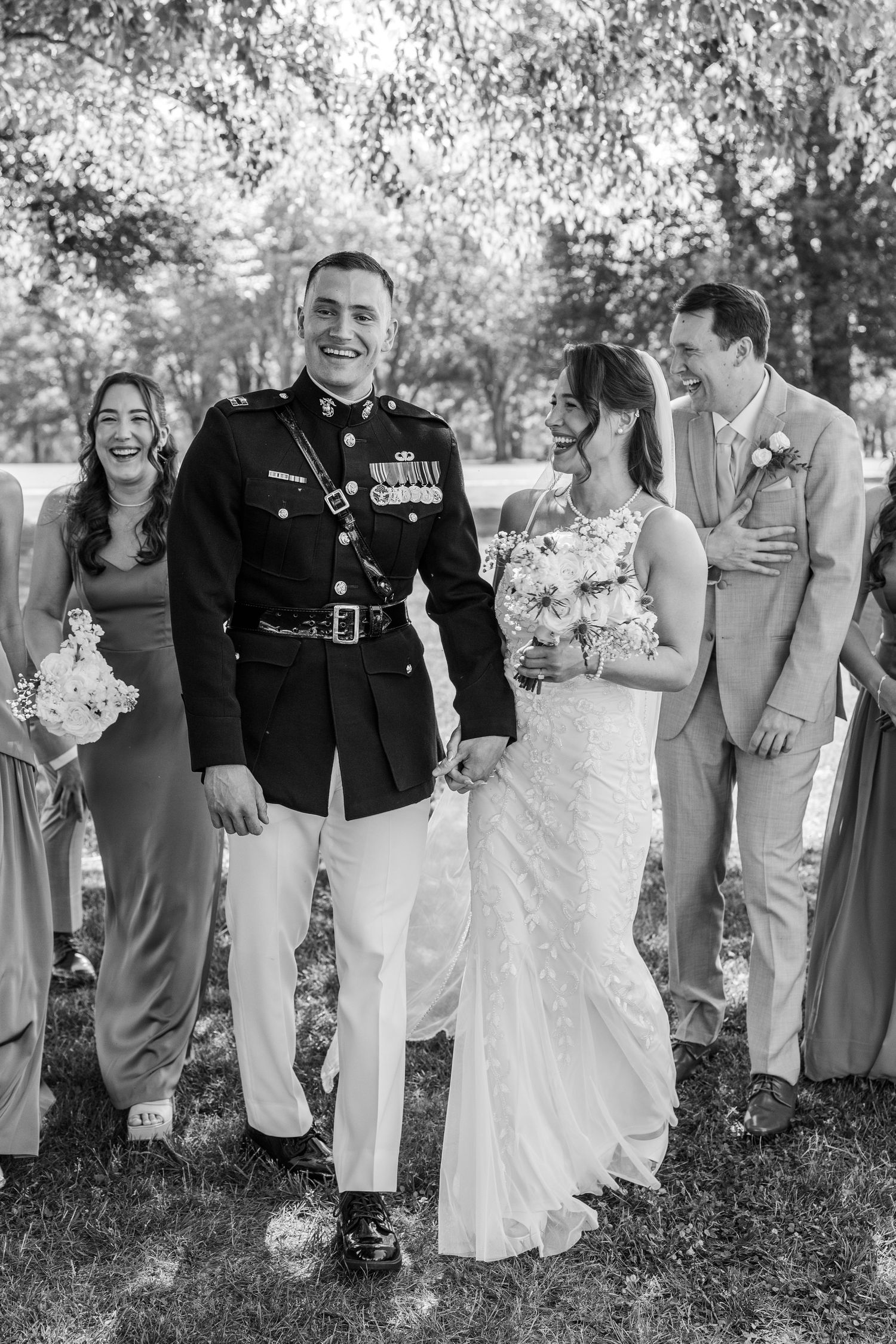 Black and white photo of a smiling military groom in dress uniform holding hands with his bride at their outdoor wedding.