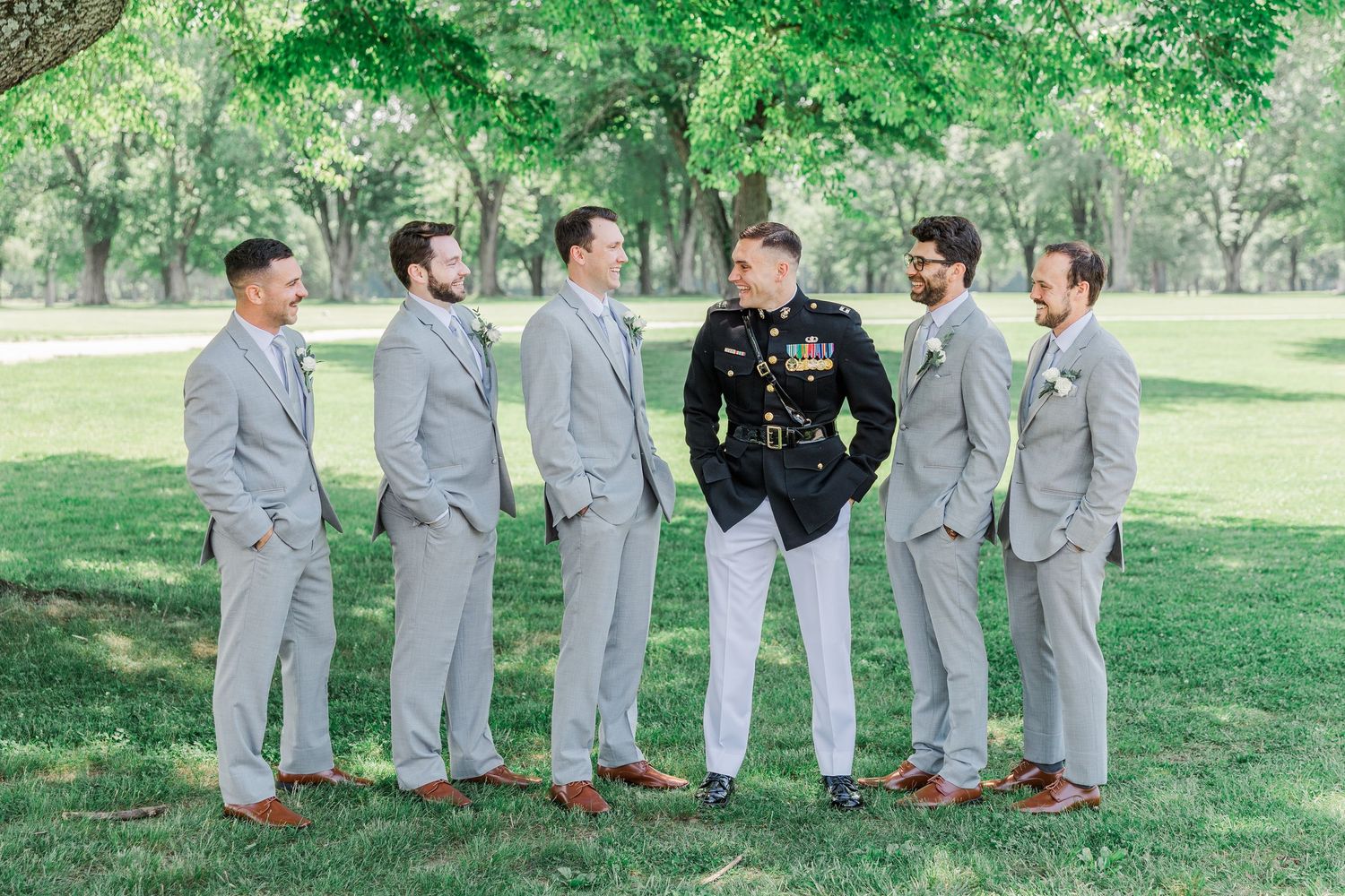 Groom in Marine dress uniform stands with five groomsmen in light gray suits on green lawn under trees.