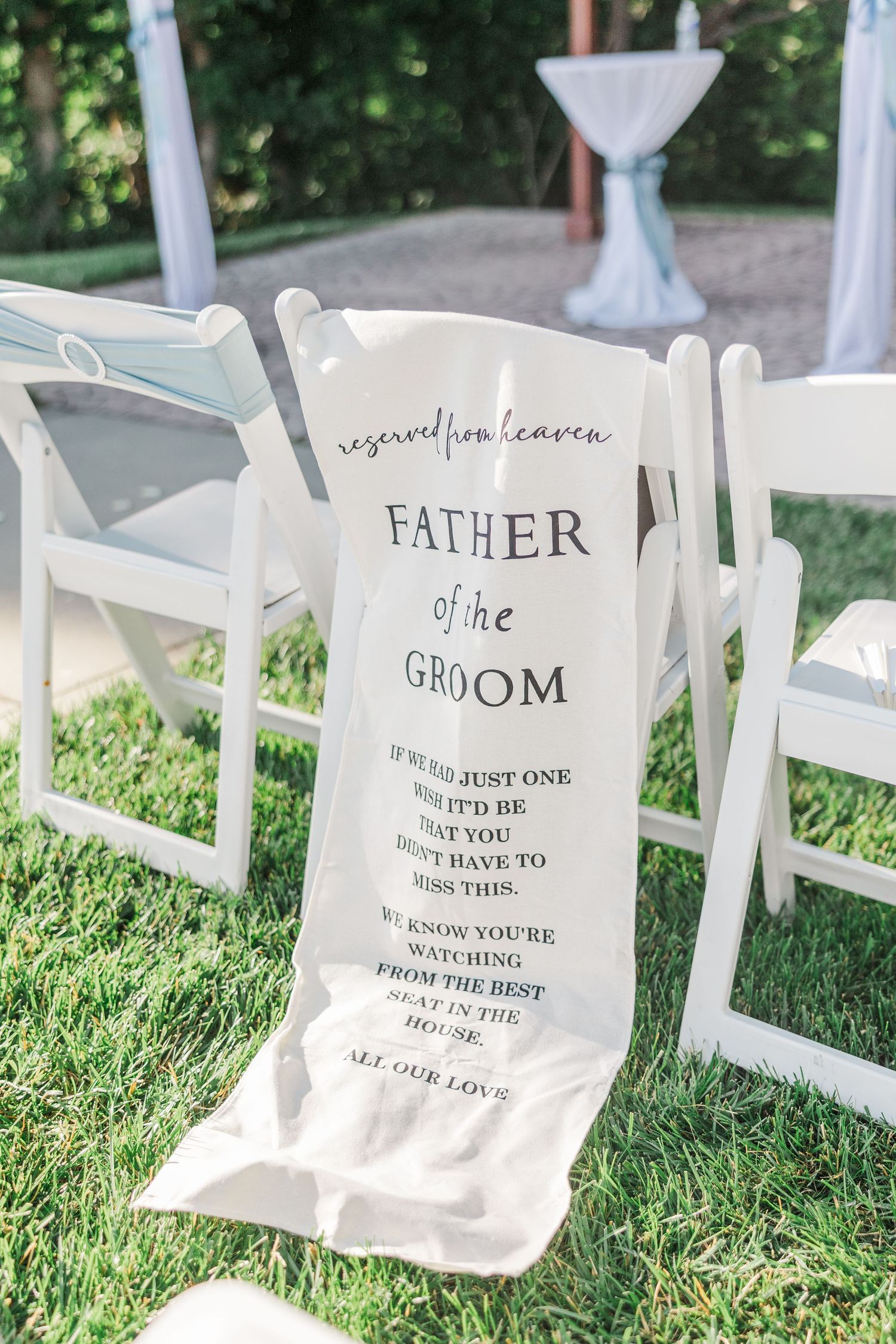 Reserved from heaven Father of the Groom memorial chair sign at an outdoor wedding ceremony on green grass.