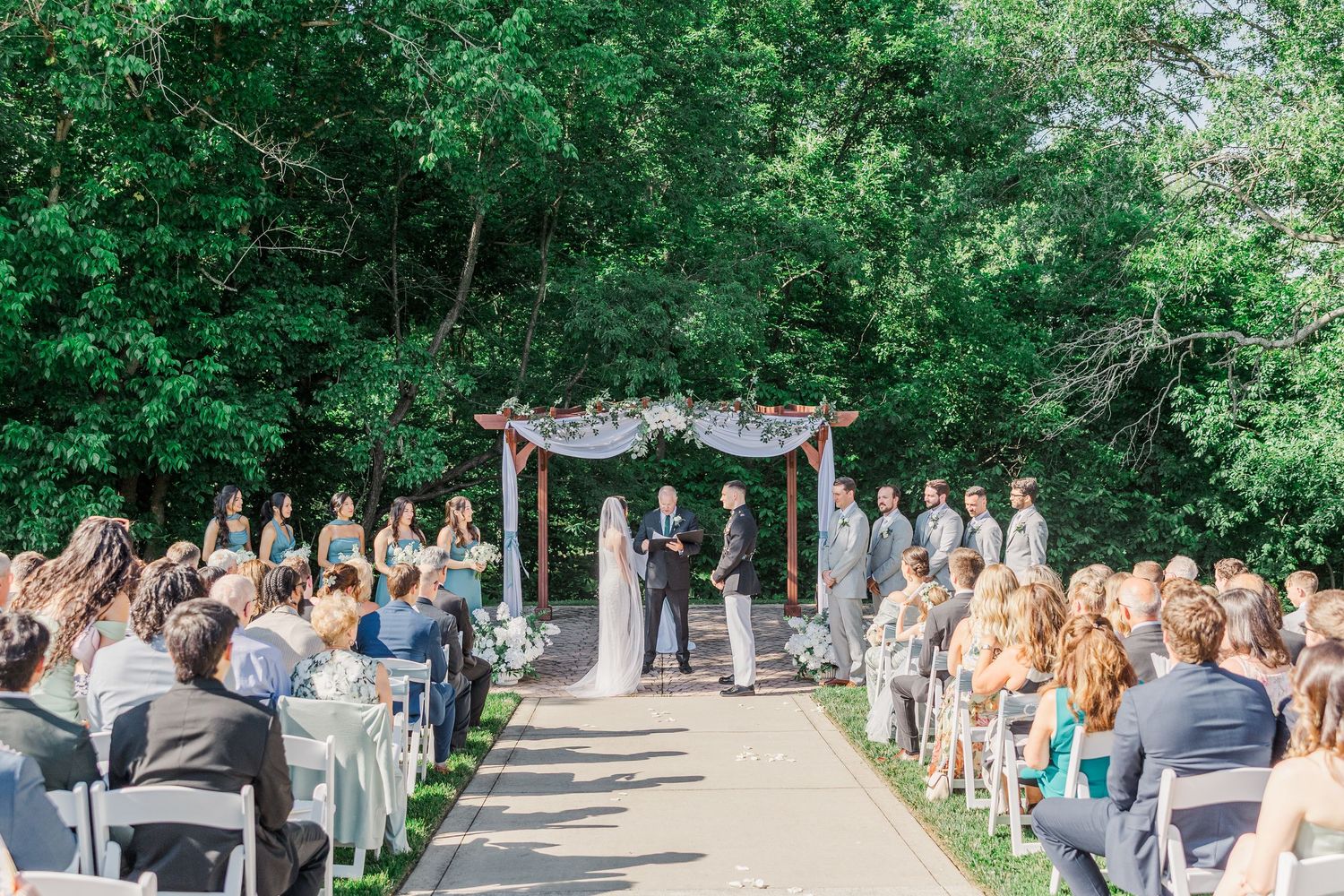 Outdoor wedding ceremony with couple at floral arch, guests seated on white chairs, surrounded by lush green trees.