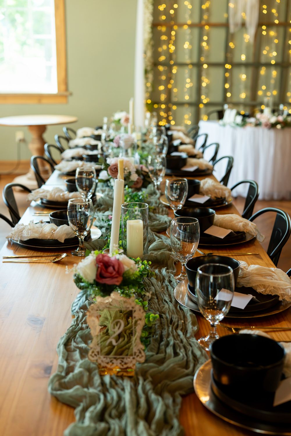 Elegant wedding reception table with green runner, black plates, candles, floral centerpieces, and fairy lights in background.