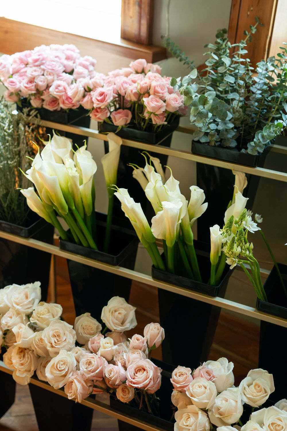 Flower shop display with pink roses, white calla lilies, and eucalyptus arranged in black vases on shelves.