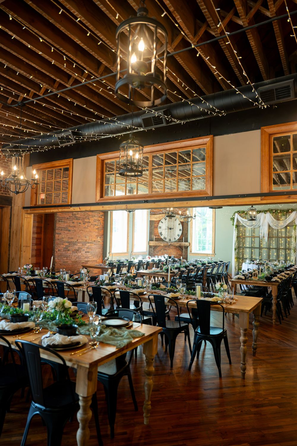 Rustic wedding reception venue with string lights, wooden tables, black chairs, brick walls, and elegant chandeliers.