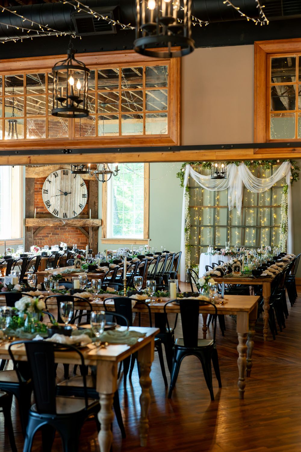 Rustic wedding reception venue with farmhouse tables, black metal chairs, Edison lights, large clock, and lush greenery decor.