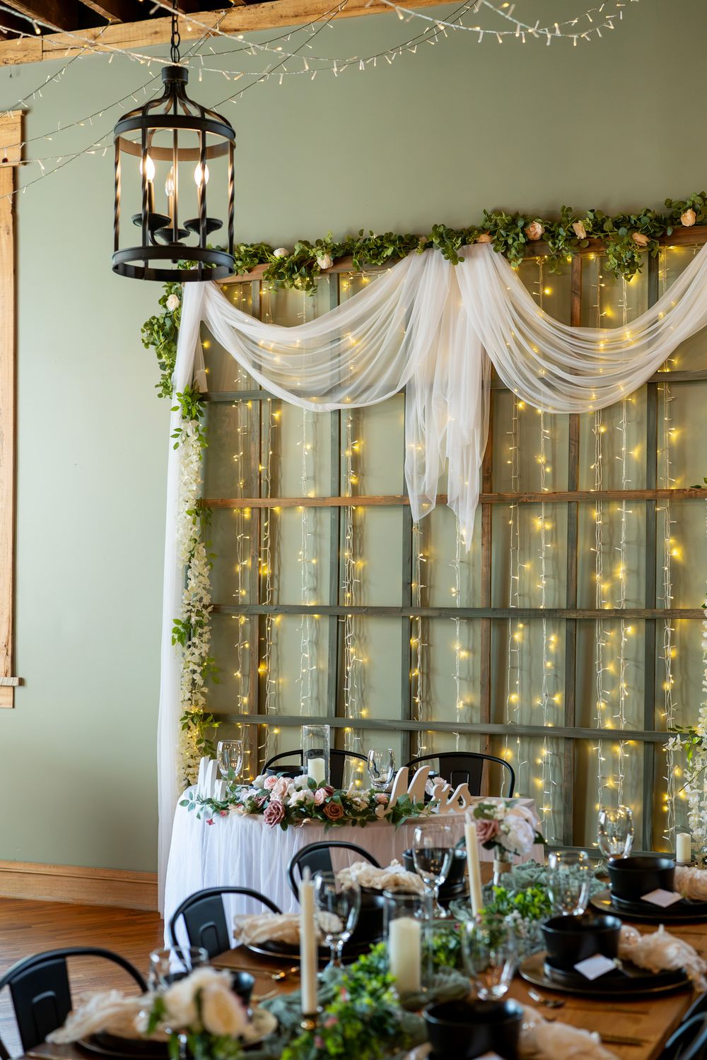 Elegant wedding reception table with greenery, candles, and fairy light backdrop with white draping.