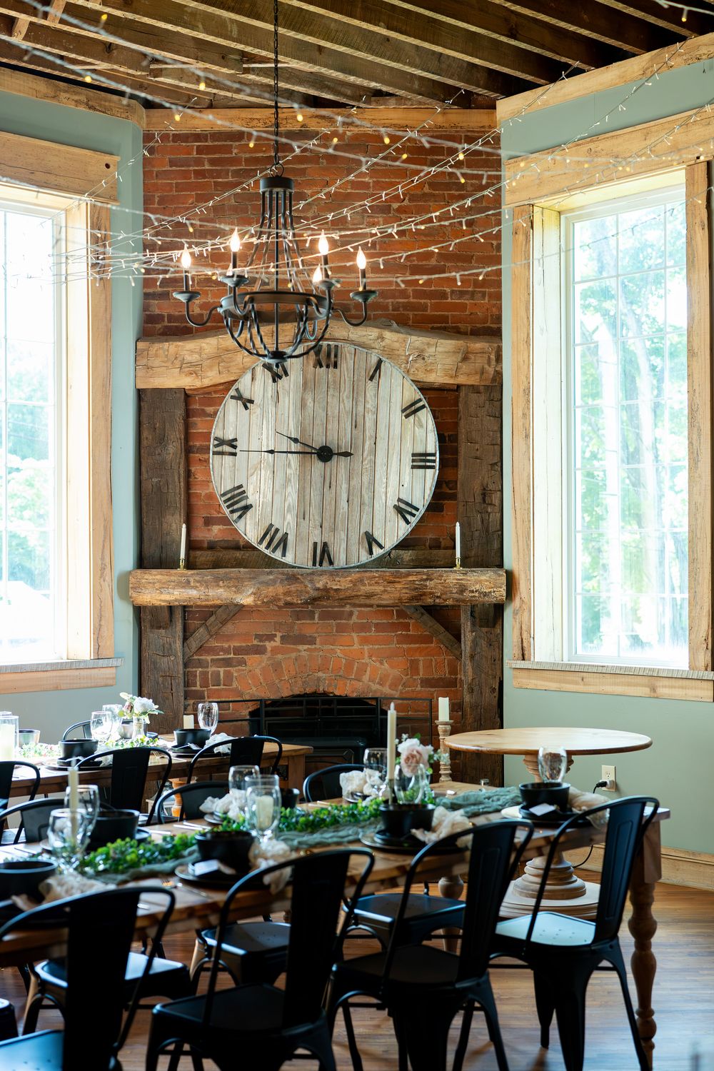 Rustic dining room with large wooden clock, brick fireplace, chandelier, string lights, and elegantly set farmhouse tables.