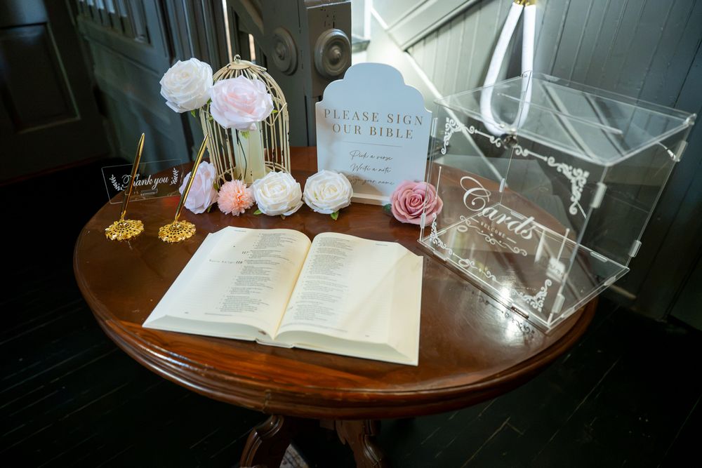 Wedding guest book table with open Bible, pink floral decor, acrylic card box, and elegant signage on wooden round table.