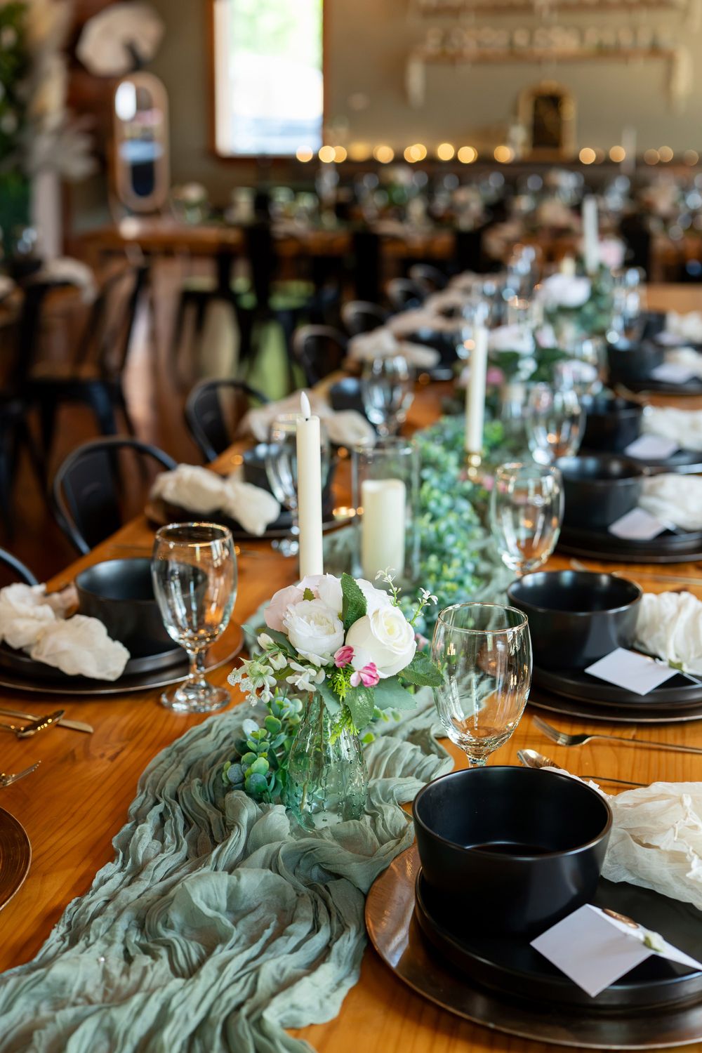 Elegant wedding reception table with black plates, white candles, greenery runner, and floral centerpieces on a wooden table.