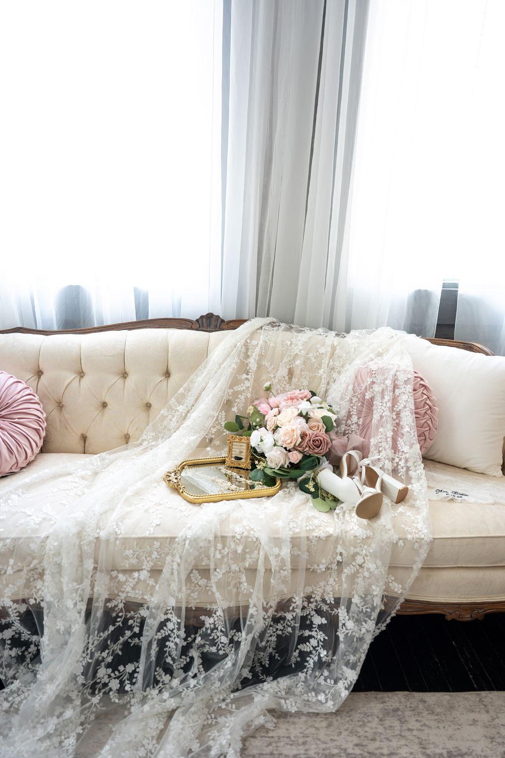 Elegant bridal flat lay with lace veil, pink rose bouquet, white heels, and gold tray on a tufted ivory sofa.
