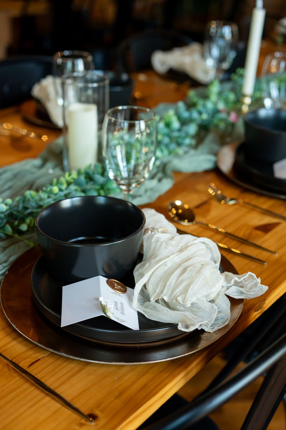 Elegant table setting with black plates, gold cutlery, white napkin, candles, and greenery on a wooden table.