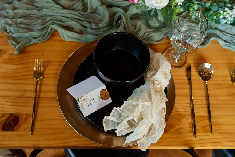 Elegant wedding table setting with black bowl, gold charger, ruffled napkin, place card, and gold cutlery on wood table.
