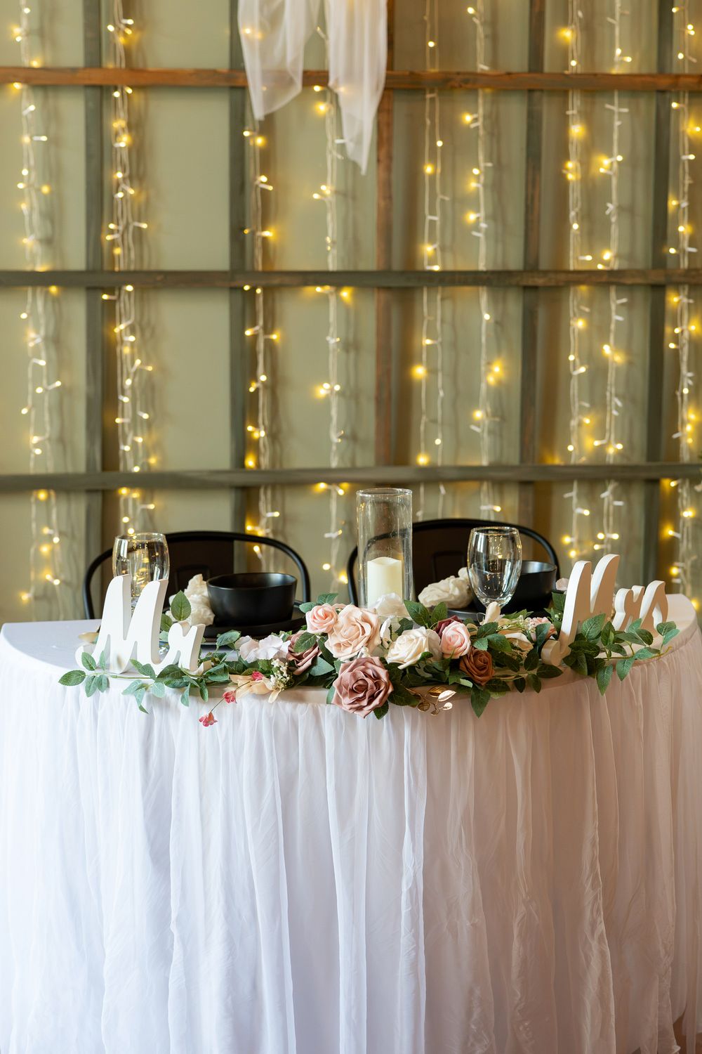 Elegant wedding sweetheart table with floral garland, candles, Mr and Mrs signs, and fairy lights backdrop.