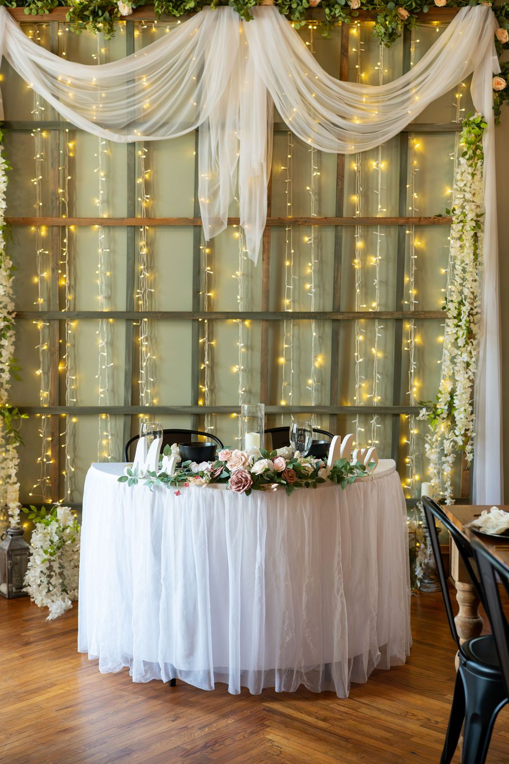 Elegant wedding sweetheart table with white tulle, floral arrangements, candles, and fairy lights backdrop in a rustic venue.