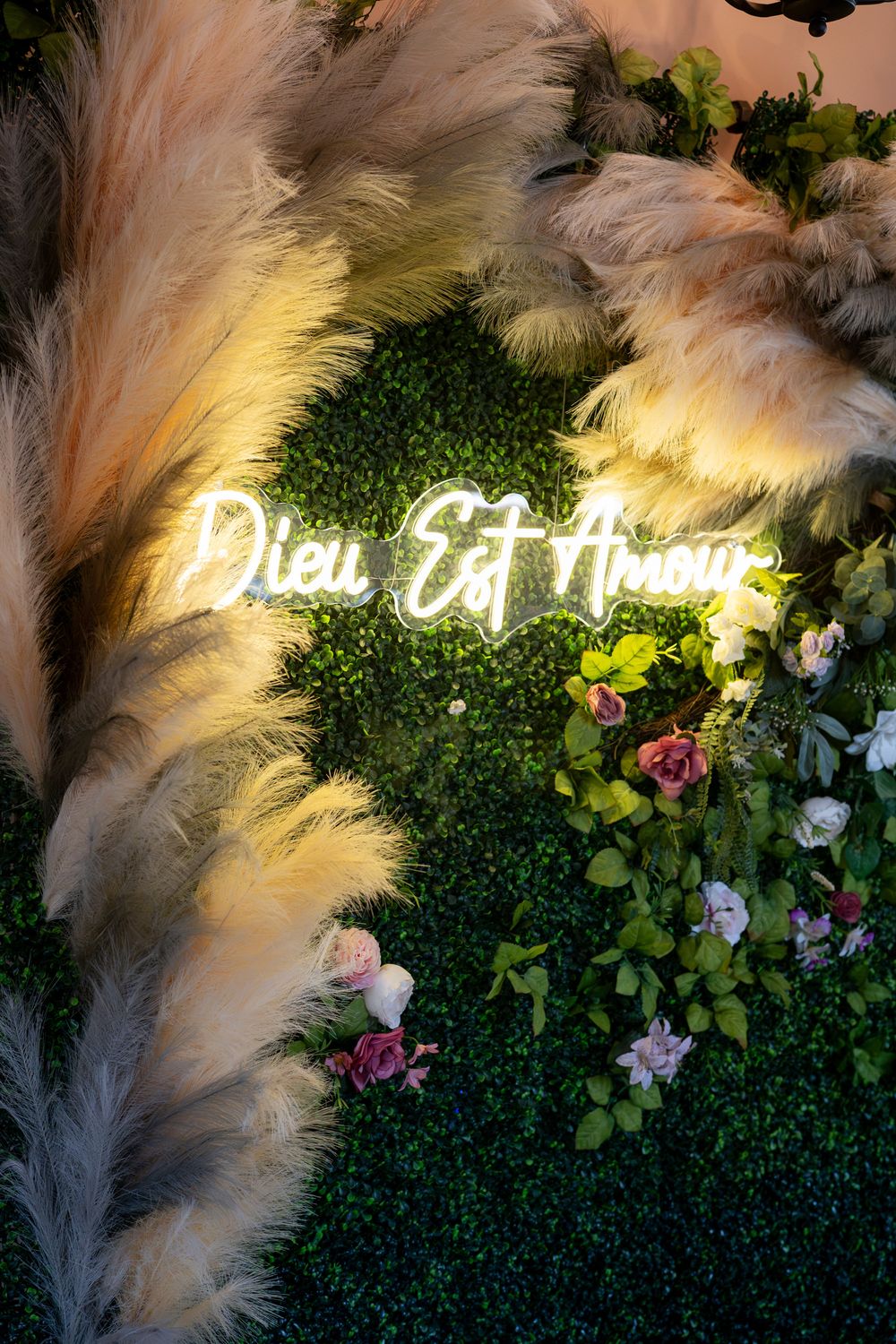 Neon sign reading Dieu Est Amour on a green floral wall with pampas grass and pink roses.