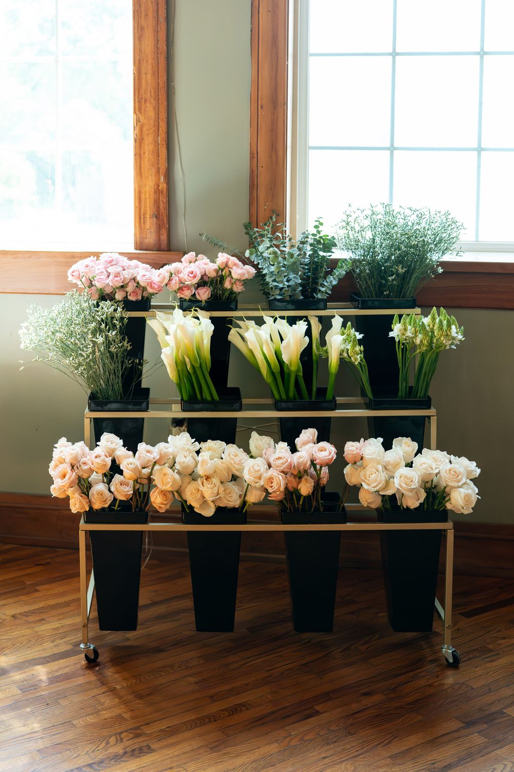 Three-tier gold rolling cart displaying fresh pink roses, white calla lilies, and greenery in black vases near a sunny window.