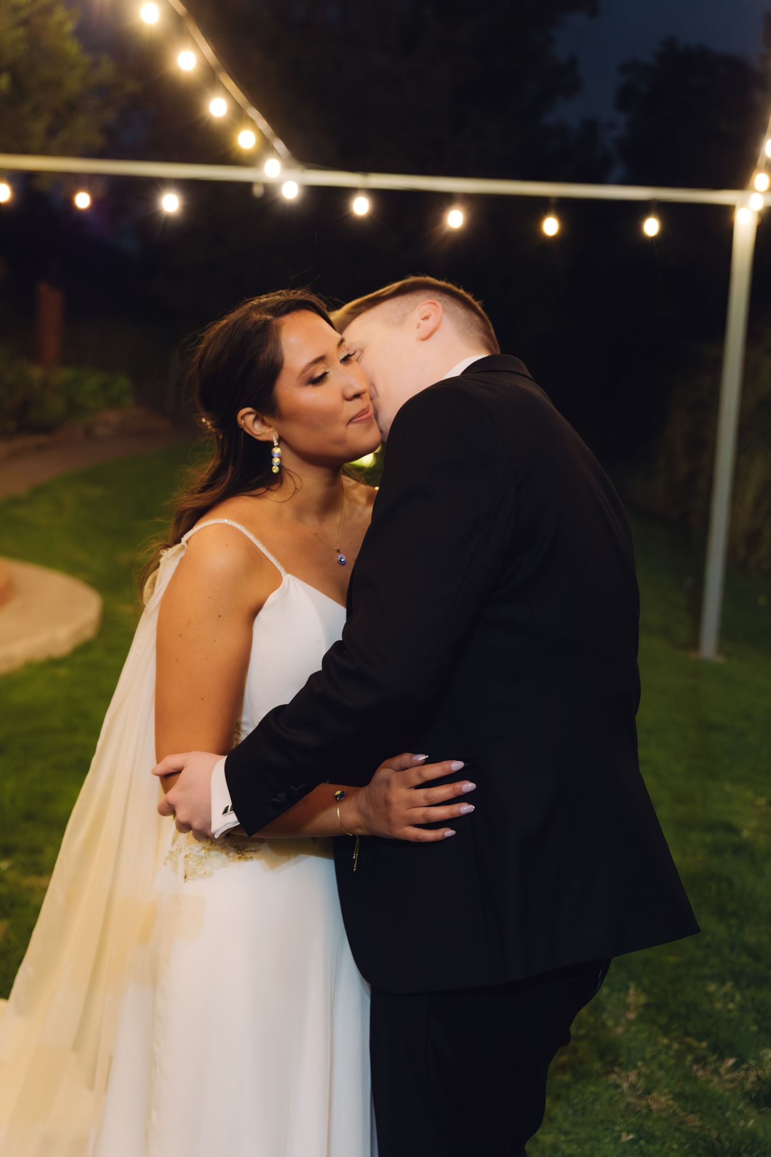 Bride and groom share a tender kiss under warm string lights during their outdoor evening wedding reception.