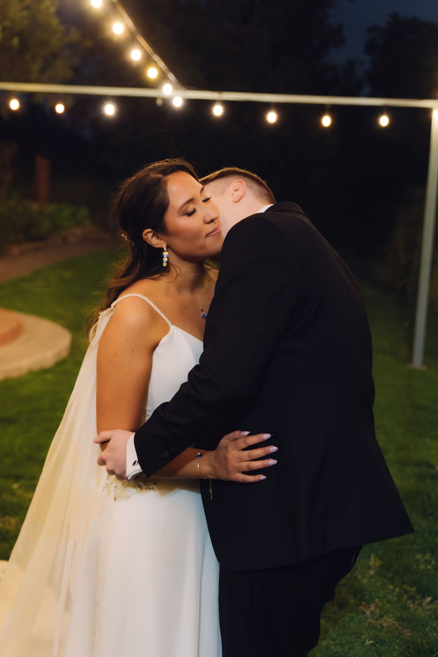 Groom kisses bride on the cheek as they embrace under glowing string lights at their outdoor evening reception.