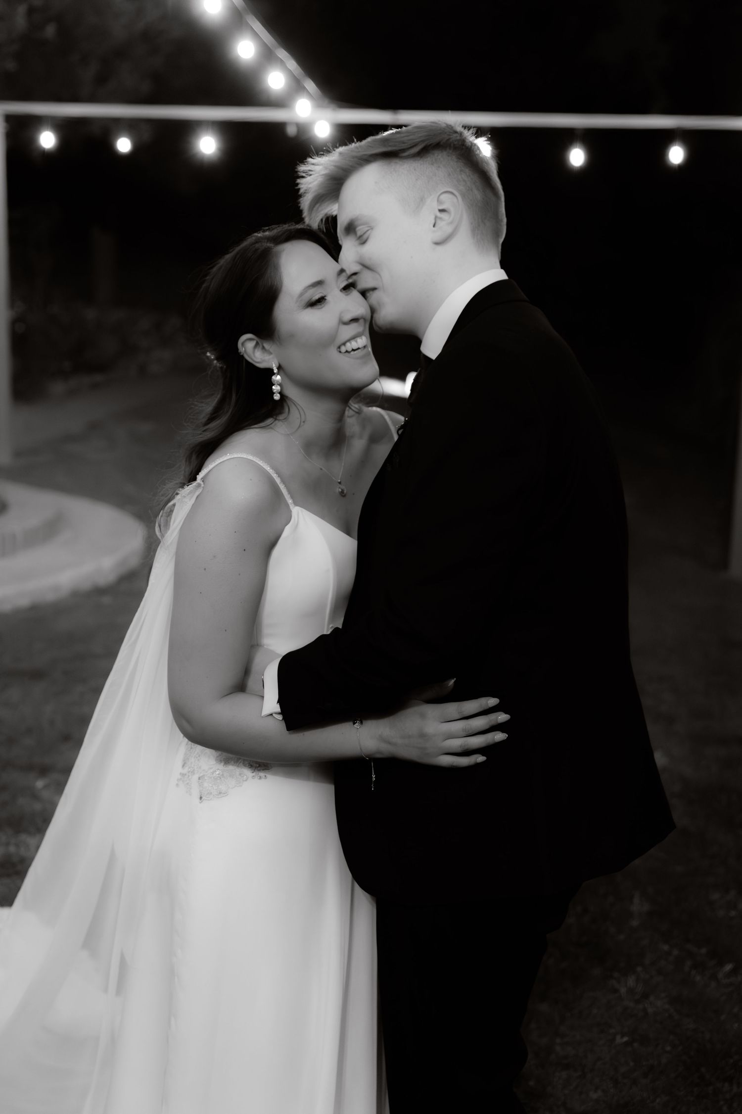 Black and white photo of smiling bride and groom sharing an intimate moment under string lights at night.