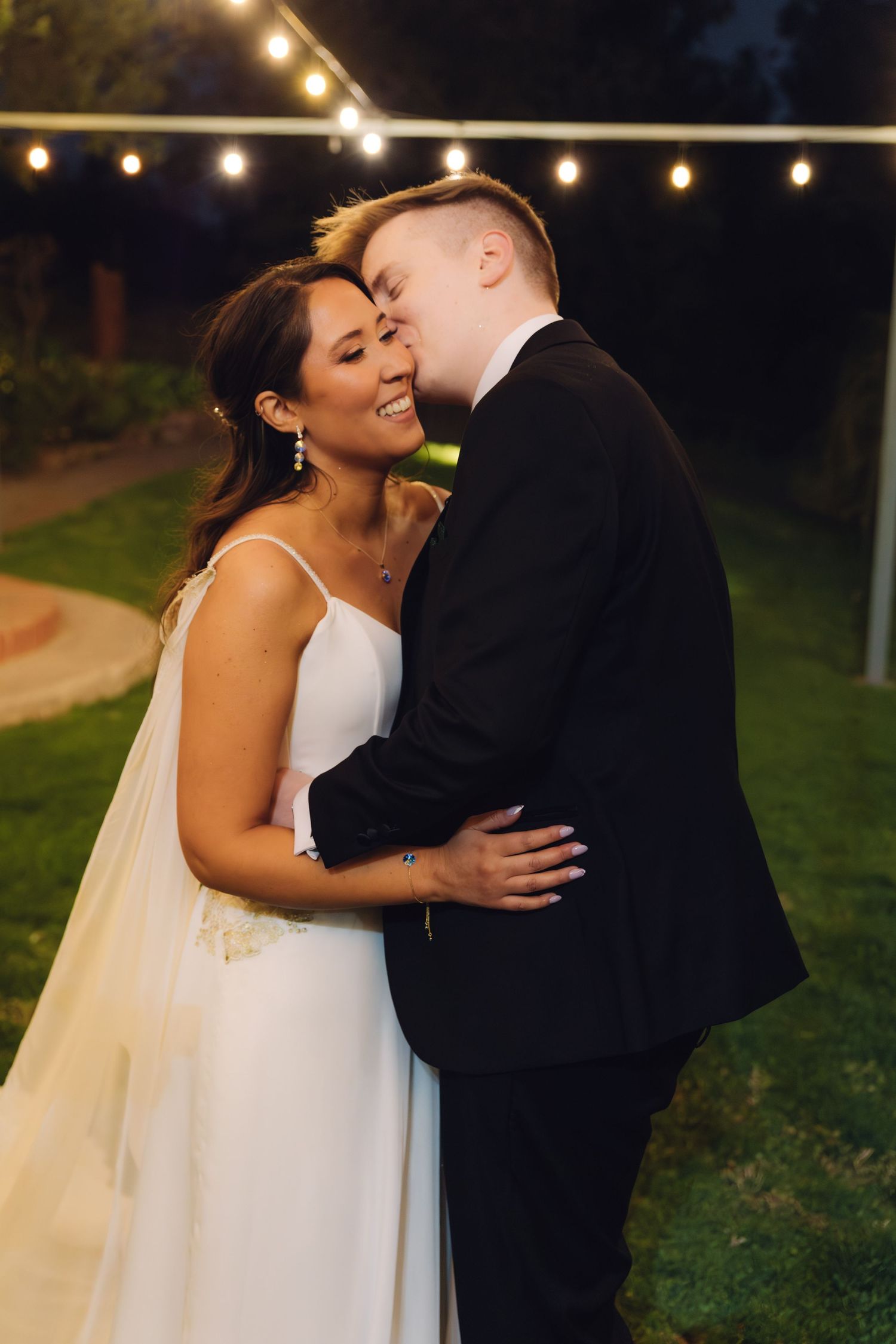 Groom kisses bride on the cheek as she smiles joyfully under string lights at their outdoor night wedding.