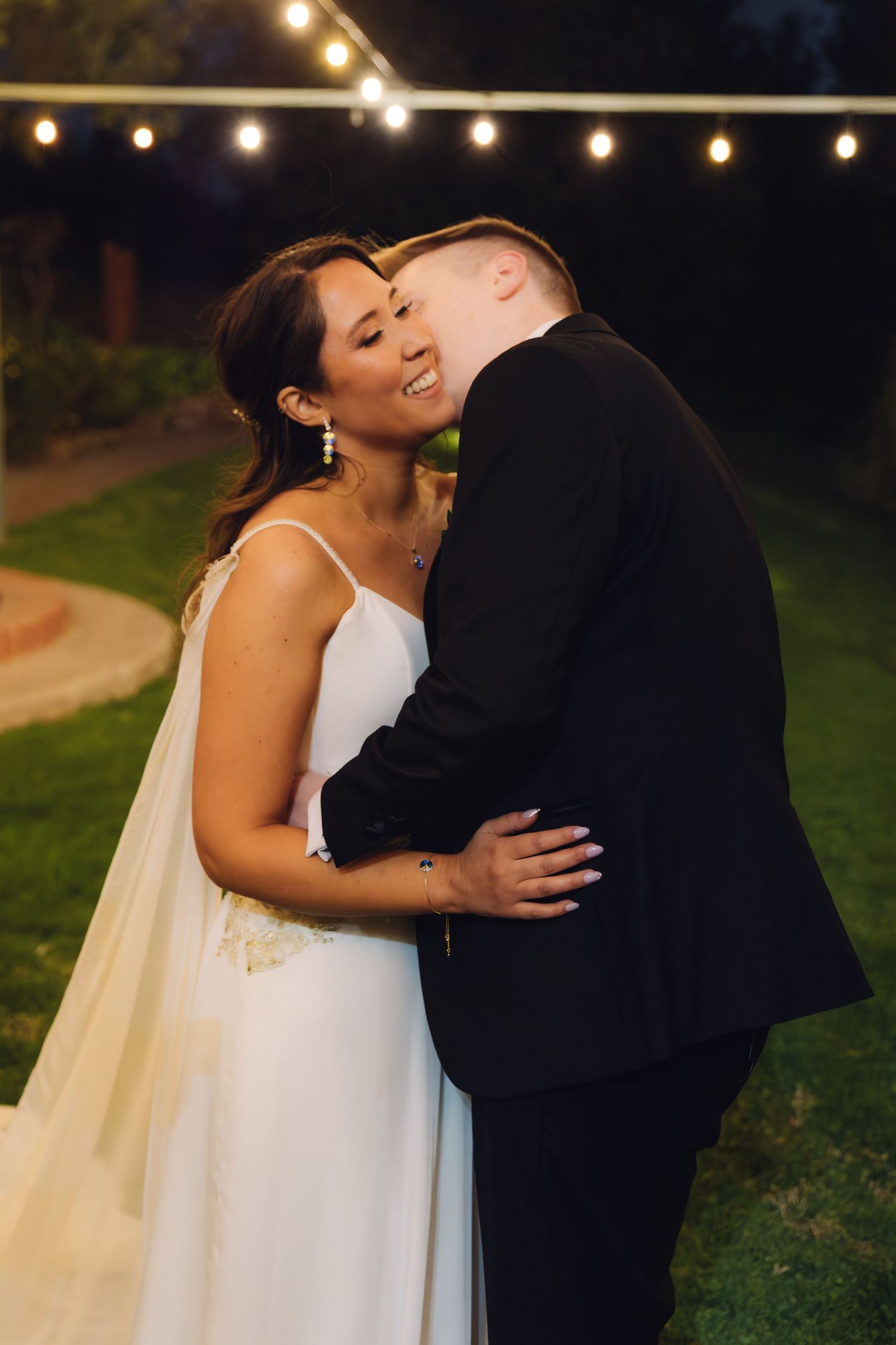 Bride smiling joyfully as groom kisses her cheek under romantic string lights at outdoor wedding.