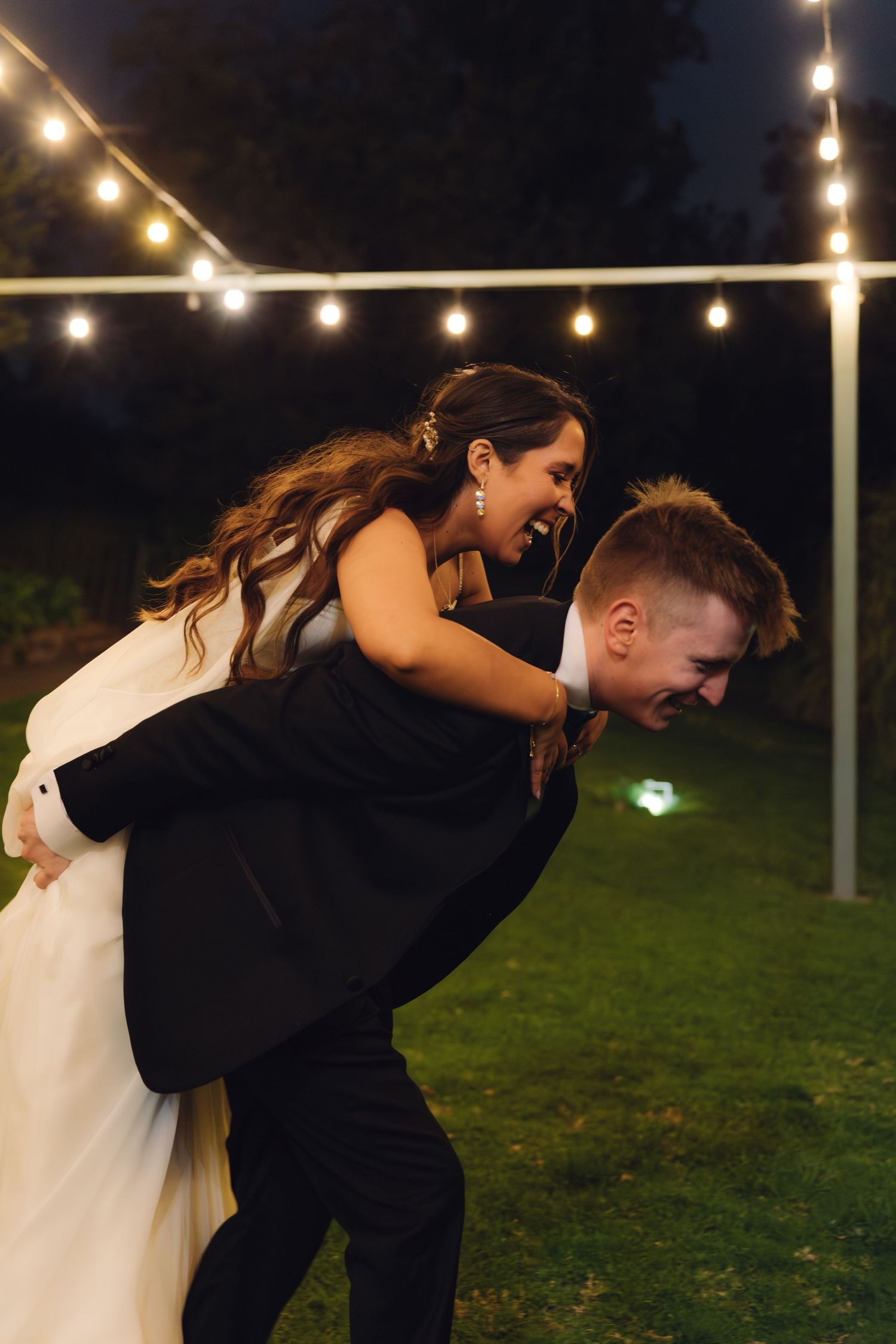 Laughing groom gives bride a playful piggyback ride under string lights at their outdoor evening wedding reception.